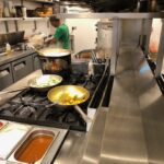 Close-up view of a commercial kitchen hot line at La Galleria, focusing on a four-burner gas hob with two frying pans containing cooking vegetables and a large pot of liquid in the background.