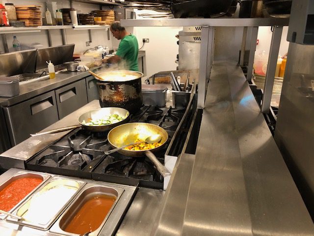 Close-up view of a commercial kitchen hot line at La Galleria, focusing on a four-burner gas hob with two frying pans containing cooking vegetables and a large pot of liquid in the background.